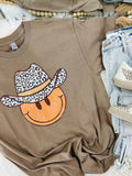 A brown short-sleeved graphic tee with a leopard wearing a cowboy hat and the text 'HAPPY' on it, displayed on a surface with jeans and shoes.