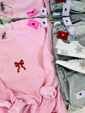 The image displays a light pink embroidered sweatshirt with the phrase "BE MINE" and a red ribbon design, presented next to a pair of blue denim jeans. The sweatshirt features decorative red heart-shaped tags and embroidered stars, enhancing its Valentine's Day-themed aesthetic.