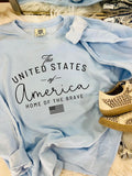 A light blue t-shirt with the text 'The United States of America Home of the Brave' printed on it, displayed flat with a pair of beige shoes at the bottom of the frame.