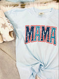 A blue curved-neck t-shirt with the word 'MAMA' and a star graphic in red and blue colors, displayed flat with a pair of white sneakers and a furry white blanket in the background.