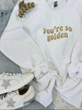 A white retro wave embroidered sweatshirt with the text 'you're so golden' on the front, displayed on a furry surface with a pair of sneakers and a star-shaped brooch.