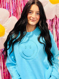 A person wearing a blue sweatshirt with a happy face embroidery on the front, posing against a pink glittery background with balloons.