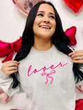 A woman smiling and posing with a gray sweatshirt that has the word 'lover' written in neon pink and a pink bow graphic.