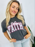 A woman wearing a gray t-shirt with the word 'Faith' and a pink bow on it, standing with one hand against a white background.