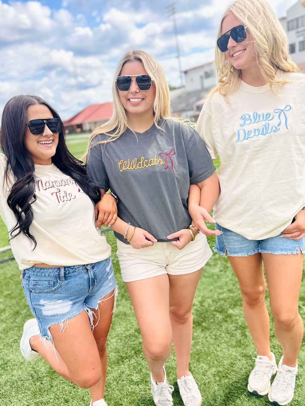Three women standing on a grassy field wearing casual outfits with visible mascot school  text on their shirts.