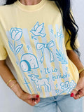 Person wearing a yellow t-shirt with blue illustrations and IT IS FINISHED text.