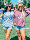 Two women standing on a field wearing sunglasses and custom mascot sweatshirts in blue and gray, with one woman's shirt displaying the word 'DEVILS' and the other's shirt displaying the word 'TIDE'.