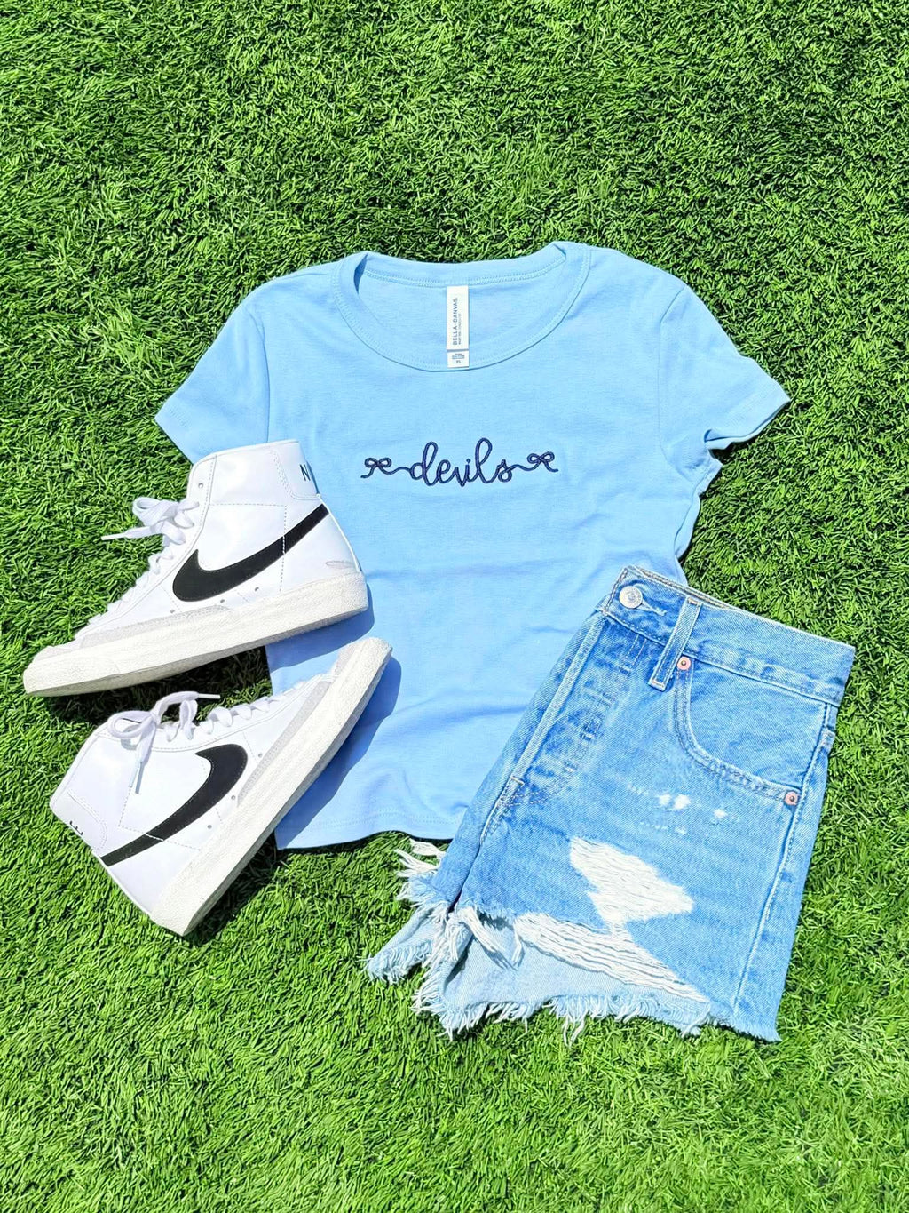 Light blue t-shirt with 'devils' text, white high-top sneakers, and denim shorts on grass