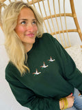 A woman sitting on a wooden chair wearing a green embroidered sweatshirt with a trio of mallard ducks on the front.