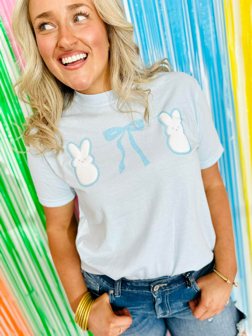 Person wearing a white t-shirt with blue designs with Puff Peeps Bunny and glitter bows against a colorful striped background