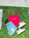 Pink tank top with text, denim shorts, sneakers, and sunglasses on grass